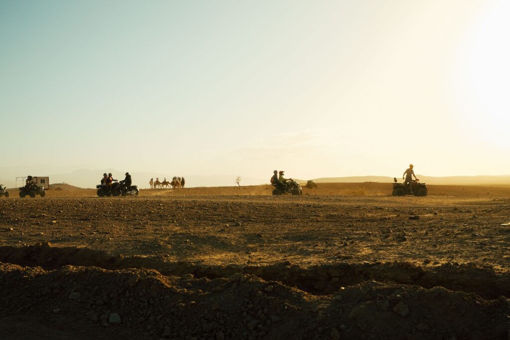 Quad bikes and camels traverse an expansive desert landscape, illuminated by warm, golden sunlight.